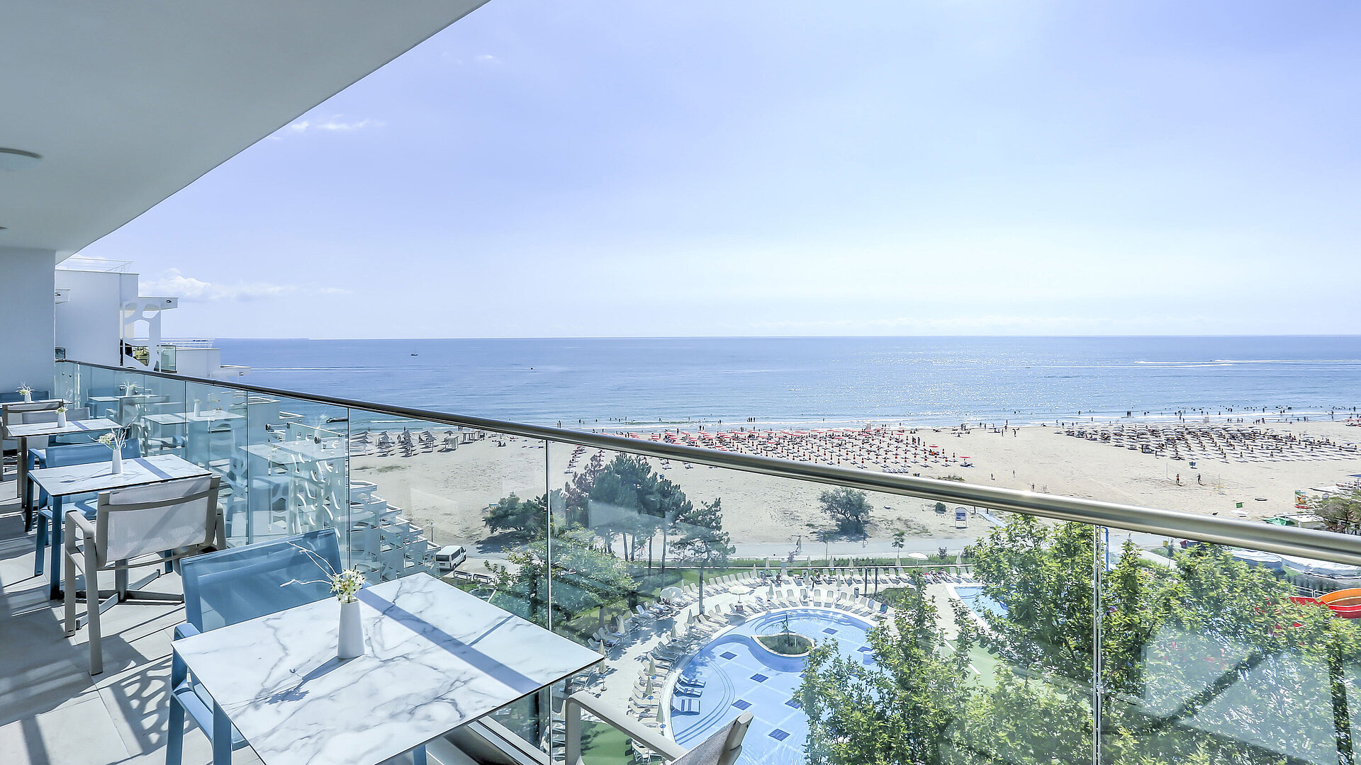 Balkonterrasse mit Tischen, Glasgeländer und weitem Blick auf Strand, Meer und Resortpools
