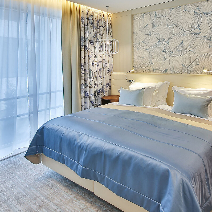 Modernes Hotelzimmer mit Doppelbett, blauen Akzenten und großen Fenstern mit Vorhängen